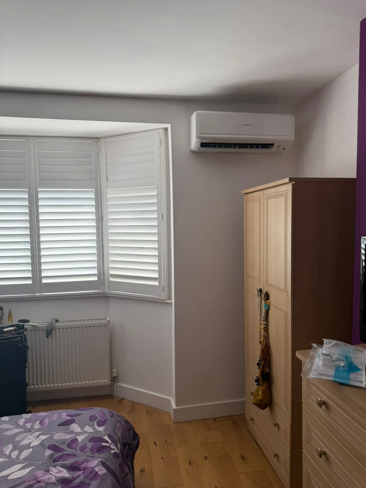 Wall-mounted air conditioning unit installed in a bedroom beside shutters